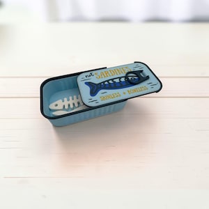May include: A light blue rectangular container with a sliding lid featuring a dark blue fish graphic, a black pull tab, and the text "not SARDINES" and "SKINLESS + BONELESS". Inside the container lies a white, skeletal fish-shaped object.