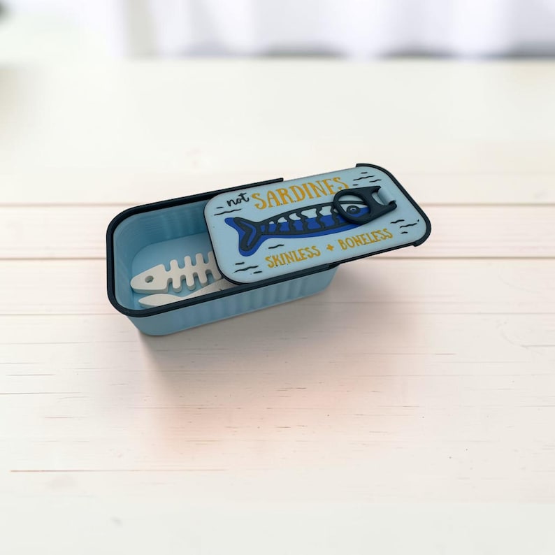 May include: A light blue rectangular container with a sliding lid featuring a dark blue fish graphic, a black pull tab, and the text "not SARDINES" and "SKINLESS + BONELESS". Inside the container lies a white, skeletal fish-shaped object.