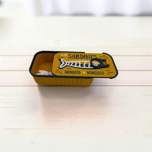 May include: A rectangular yellow container with a dark blue rim, featuring a lid printed with a stylized fish graphic and the text "not SARDINES" and "SKINLESS + BONELESS". The lid includes a dark blue pull-tab design on the top surface.