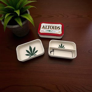 May include: A red and white Altoids mint tin sits above two white rectangular trays. One tray has a green cannabis leaf design, and the other has a cannabis leaf and a black and white graphic. A potted plant is in the background.