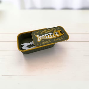 May include: An olive green rectangular container with a sliding lid featuring a yellow graphic of a fish skeleton and the text "not SARDINES" along with "SKINLESS" and "BONELESS". Inside the container rests a white fish skeleton cutout.