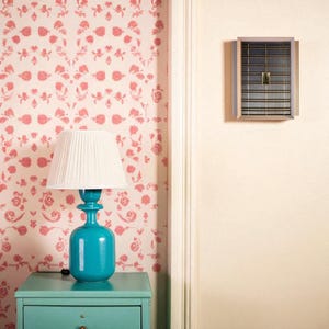 May include: A teal bedside table with a matching lamp. The lamp has a white pleated shade and a teal base. The wallpaper is a light pink with a floral pattern. A rectangular vent is mounted on the wall.