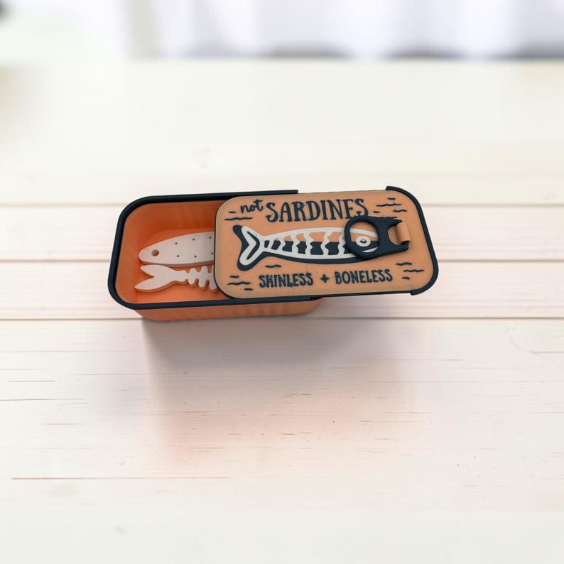 May include: A rectangular orange container with a black rim and a sliding lid. The lid features a graphic of a fish skeleton and the text "not SARDINES" and "SKINLESS + BONELESS". A white fish skeleton shape is visible inside the open container.