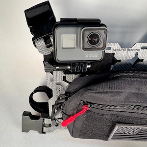 May include: A black tactical chest rig with a GoPro camera mounted on the front. The rig has a small black pouch with a red zipper pull and a gray and black American flag patch.