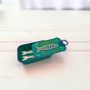 May include: A rectangular teal container with a lid shaped like a sardine tin. The lid features a blue and gold fish graphic with the text "not SARDINES" and "SKINLESS BONELESS". Inside the container are two white fish-shaped objects.