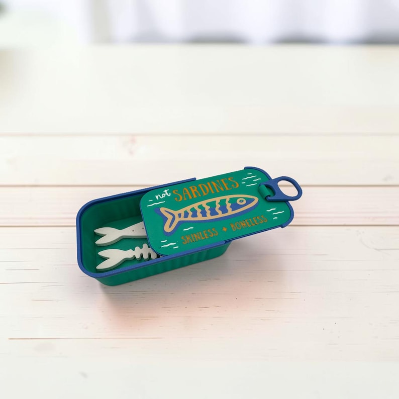 May include: A rectangular teal container with a lid shaped like a sardine tin. The lid features a blue and gold fish graphic with the text "not SARDINES" and "SKINLESS BONELESS". Inside the container are two white fish-shaped objects.