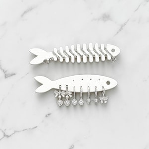 May include: Two silver-toned fish-shaped jewelry organizers. The top fish has a textured spine for hanging items. The bottom fish features holes for displaying six dangling charms, including bows with hearts, round stones, starbursts, and a butterfly.