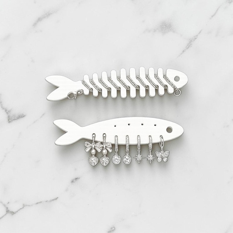 May include: Two silver-toned fish-shaped jewelry organizers. The top fish has a textured spine for hanging items. The bottom fish features holes for displaying six dangling charms, including bows with hearts, round stones, starbursts, and a butterfly.