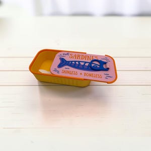 May include: A rectangular yellow container with an orange rim and a sliding lid. The lid is light purple with a blue fish graphic, a blue pull-tab, and the text "not SARDINES" and "SKINLESS + BONELESS" printed in orange and dark blue.