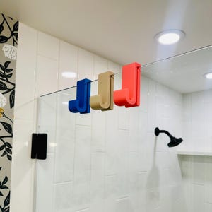 May include: Three colorful shower door hooks in blue, tan, and red are attached to a glass shower door. The hooks are in a bathroom with white tile and a black shower head. The hooks are designed to hang items.