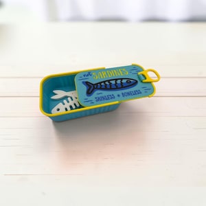 May include: A rectangular blue container with a yellow rim and a hinged lid. The lid features a graphic of a blue fish and the text "not SARDINES SKINLESS + BONELESS". Inside the container, a white fish skeleton shape is visible.