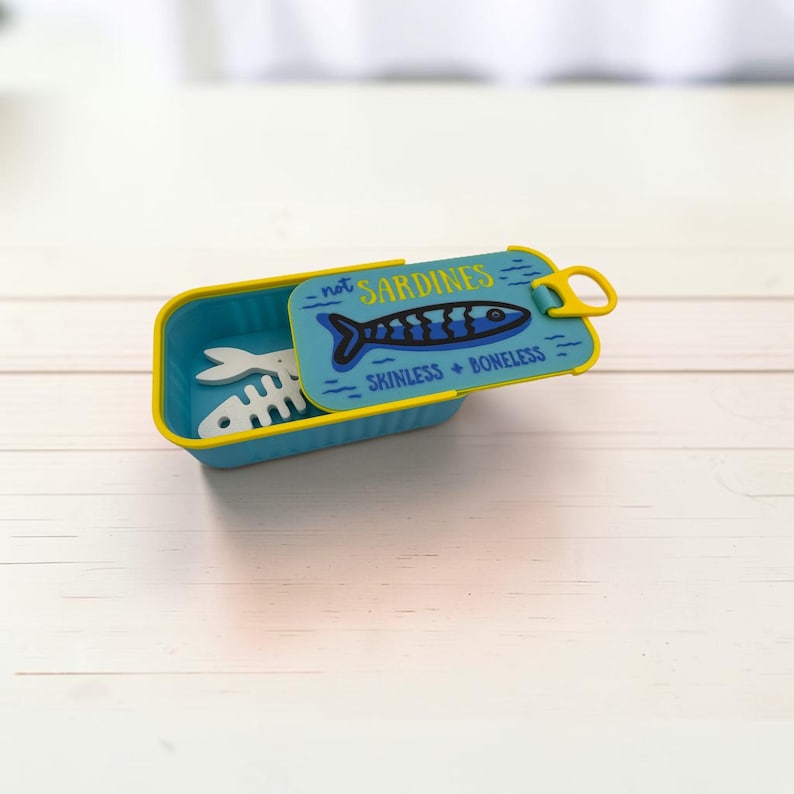 May include: A rectangular blue container with a yellow rim and a hinged lid. The lid features a graphic of a blue fish and the text "not SARDINES SKINLESS + BONELESS". Inside the container, a white fish skeleton shape is visible.