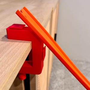 May include: A red plastic clamp with an orange plastic track attached to it. The clamp is attached to a wooden surface.