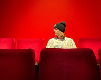 Könnte beinhalten: Eine Person mit einer schwarzen Mütze sitzt in einer Reihe roter Theaterstühle. Die Sitze sind leer, bis auf den, auf dem die Person sitzt.