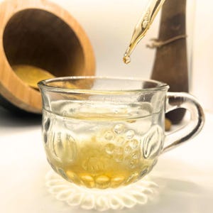 May include: A clear glass teacup with a handle, filled with a yellow liquid. A dropper dispenses a golden liquid into the cup. A wooden mortar and pestle are in the background, suggesting herbal tea or a similar concoction.
