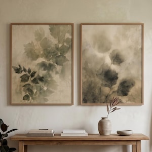 May include: Two rectangular framed wall art prints featuring soft, muted green and beige watercolor-style botanical leaf illustrations. The prints are displayed side-by-side in light-toned wooden frames, showcasing organic foliage patterns.