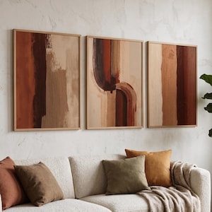 May include: Three framed abstract art pieces with warm earth-tone brushstrokes hang above a white sofa with throw pillows. A wooden coffee table displays decor and an open book. A potted plant adds greenery to the room.