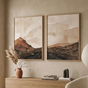 Può includere: Due stampe artistiche incorniciate con un tema montano in tonalità neutre di marrone e beige. L'opera d'arte è appesa sopra un mobile in legno con un vaso di piante secche. Un tappeto e una sedia di colore neutro completano la stanza.