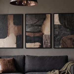 Può includere: Tre quadri rettangolari da parete con pittura a impasto spessa e strutturata nei toni del nero, marrone scuro e beige. Ogni pezzo è incorniciato da un sottile bordo nero e presenta forme geometriche astratte e sovrapposte.