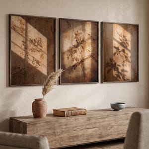 Könnte beinhalten: Drei gerahmte botanische Drucke in Braun- und Beigetönen hängen über einer Holzkonsolentisch. Die Drucke zeigen Pflanzensilhouetten und Schatten. Eine Vase mit getrocknetem Gras, ein Buch und eine Schale befinden sich auf der Konsole.