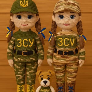 Może przedstawiać: Dwie szydełkowe lalki ubrane w kamuflażowe mundury wojskowe z żółtymi haftowanymi napisami "ЗСУ" i "ЗСВ". Lalki noszą żółte czapki z haftowanymi ukraińskimi symbolami. Mały szydełkowy pies w zielonej koszulce z haftowanym napisem "ПАТРОН" stoi między lalkami.