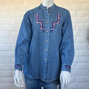 May include: A medium-wash blue denim button-down shirt featuring a mandarin collar, long sleeves, and vertical pintuck pleats. The front chest and cuffs are decorated with a geometric embroidered pattern in shades of blue, pink, and purple.