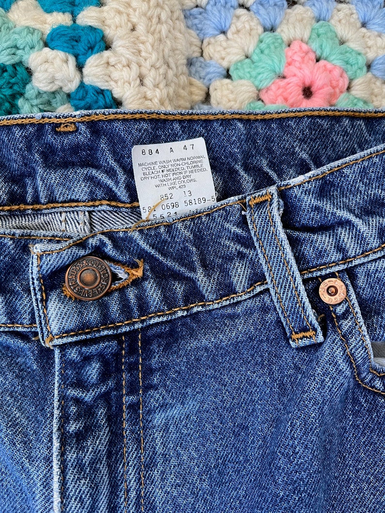 May include: Close-up of the waistband of a pair of blue denim jeans with a care label attached. The label reads "Machine wash warm normal cycle only non-chlorine bleach, tumble dry hot, hot iron if needed, wash and dry with like colors, WPL 423, 852 13, 581 0698 58189-5, 552".