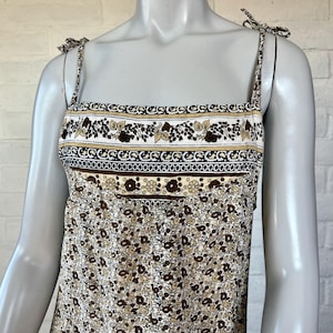 May include: A floral print sundress with adjustable tie straps. The dress features a square neckline and a bodice with a decorative band of brown, tan, and white floral and geometric patterns. The skirt has a repeating pattern of small brown flowers on a cream background.