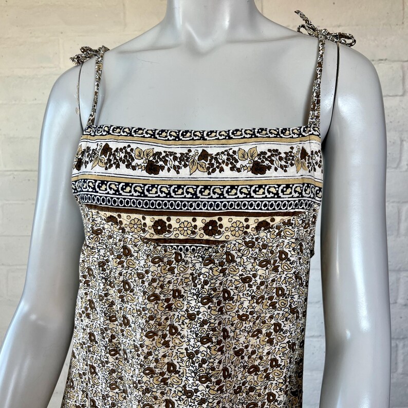May include: A floral print sundress with adjustable tie straps. The dress features a square neckline and a bodice with a decorative band of brown, tan, and white floral and geometric patterns. The skirt has a repeating pattern of small brown flowers on a cream background.
