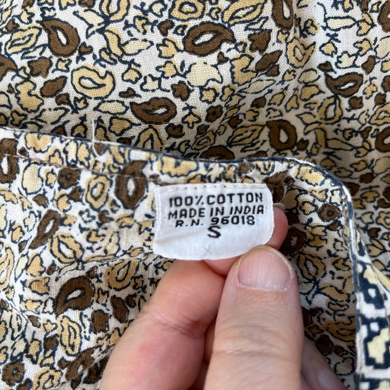 May include: Close-up of a floral patterned fabric in cream, brown, and yellow. The fabric has a small white tag with the text "100% Cotton Made in India R.N. 96018 S".