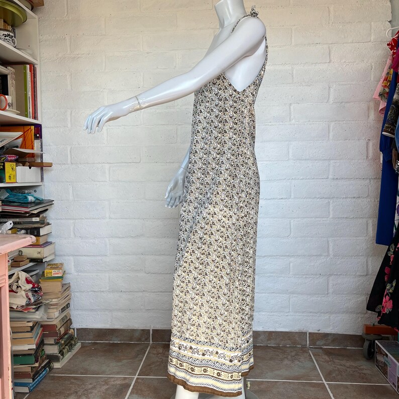 May include: A floral print maxi dress with a V-neck and spaghetti straps. The dress features a cream-colored background with small brown and black floral patterns. The hemline has a decorative border with brown and blue accents. The dress is displayed on a mannequin.