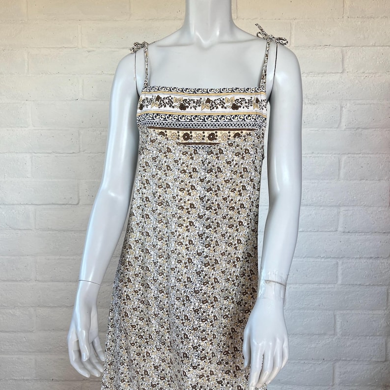 May include: A floral print dress with adjustable tie straps. The dress features a square neckline and a detailed band across the chest with floral and geometric patterns in brown, white, and blue. The dress has a light beige background with small brown floral patterns.