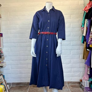 May include: A solid navy blue midi-length shirt dress featuring a classic collar, a full-length front button placket, and three-quarter length sleeves. The dress is cinched at the waist with a thin, woven red belt featuring a gold-toned buckle.
