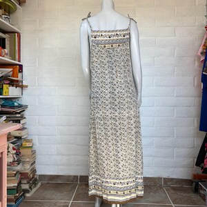May include: A long, floral-print dress with adjustable spaghetti straps. The dress features a cream-colored background with a small, all-over floral pattern in shades of blue and brown. The bodice and hemline are accented with patterned bands.