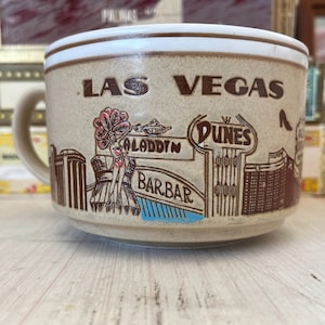 May include: A beige ceramic mug with a white rim and a brown handle. The mug is decorated with brown illustrations of Las Vegas landmarks, including the Aladdin, Dunes, and Barbary Coast. The text "LAS VEGAS" is printed on the mug.