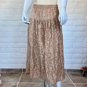 May include: A long, tan lace skirt with an elastic waistband. The skirt is displayed on a mannequin and has a floral pattern. The skirt is knee-length and has a flowing, A-line silhouette.