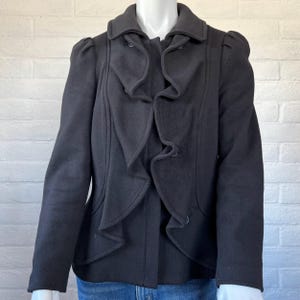 May include: A black jacket with a ruffled front and a classic collar. The jacket has a double-breasted design with two rows of buttons and features puffed sleeves. The jacket is displayed on a white mannequin against a white brick wall.