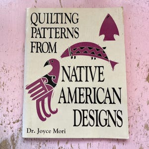 May include: A vintage book titled "Quilting Patterns From Native American Designs" with illustrations of a bird, fish, and arrowhead in shades of purple and black. The book has a cream-colored cover and is on a pink surface.