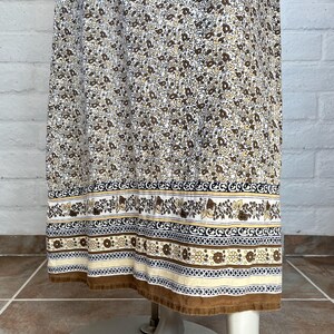 May include: A vintage-style dress with a floral pattern in shades of brown and cream. The dress features a detailed border with floral and geometric designs in brown, black, and cream. The hemline is finished with a brown trim.
