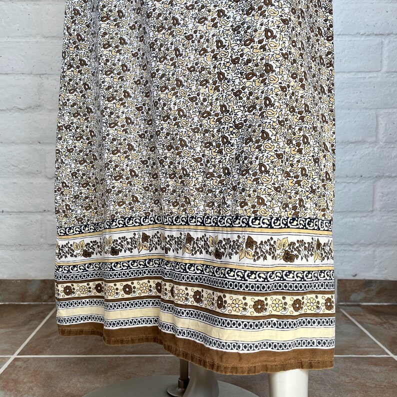 May include: A vintage-style dress with a floral pattern in shades of brown and cream. The dress features a detailed border with floral and geometric designs in brown, black, and cream. The hemline is finished with a brown trim.
