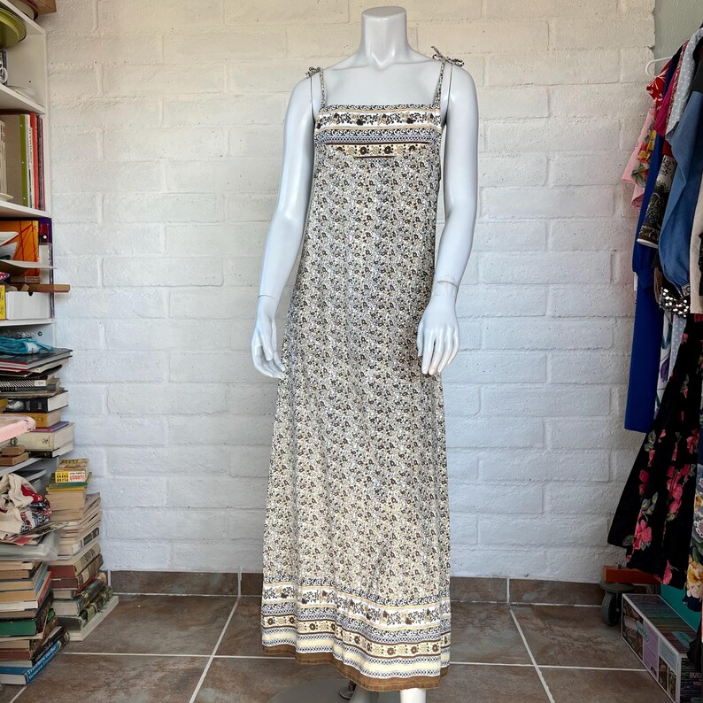May include: A long, floral-print dress on a mannequin. The dress features a cream-colored background with small blue and brown floral patterns. It has thin straps tied at the shoulders and decorative bands at the top and hem. The dress is floor-length.