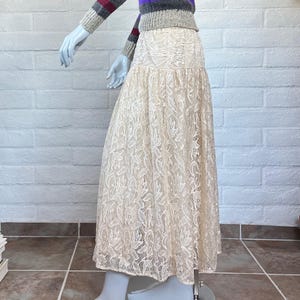 May include: A cream-colored lace skirt displayed on a mannequin. The skirt has a floral pattern and a flowing silhouette. The mannequin is also wearing a striped sweater with gray, purple, and burgundy stripes.