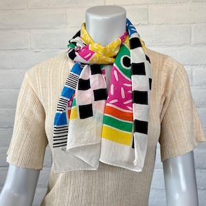 May include: A rectangular scarf featuring a vibrant, multi-colored graphic print on a white background. The design includes black and white checkerboard patterns, bright yellow, pink, green, and blue geometric shapes, and thin black stripes.