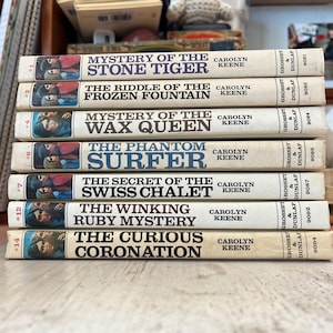 May include: A stack of vintage hardcover books featuring titles like "Mystery of the Stone Tiger" and "The Phantom Surfer." The books have colorful cover illustrations and are by Carolyn Keene. The spines are off-white with black text.