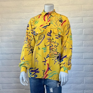 May include: Yellow button-down shirt with a vibrant, abstract print featuring giraffes, fish, and other shapes in red, blue, purple, and green. The long-sleeved shirt has two chest pockets and is displayed on a mannequin.