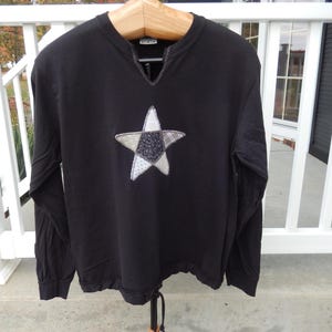 May include: Black long-sleeved shirt with a patchwork star design on the front. The star is made of different fabrics in shades of gray and white.