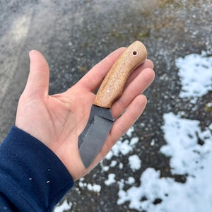 Puede incluir: Un pequeño cuchillo artesanal con mango de madera y una hoja oscura y curvada. El mango es de color marrón claro con veta de madera visible. La hoja es de color gris oscuro. El cuchillo se sostiene en una mano abierta sobre un fondo nevado.