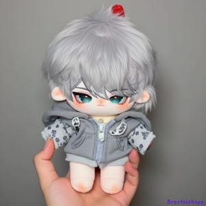 May include: A gray plush doll with silver hair, wearing a gray hooded sweatshirt with a white and gray pattern. The doll has blue eyes and a red detail on its head.