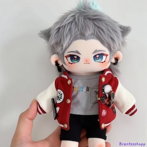 May include: A gray plush doll with a white and red varsity jacket, gray t-shirt with colorful text, black shorts, and gray hair. The doll has blue eyes and is wearing earrings.
