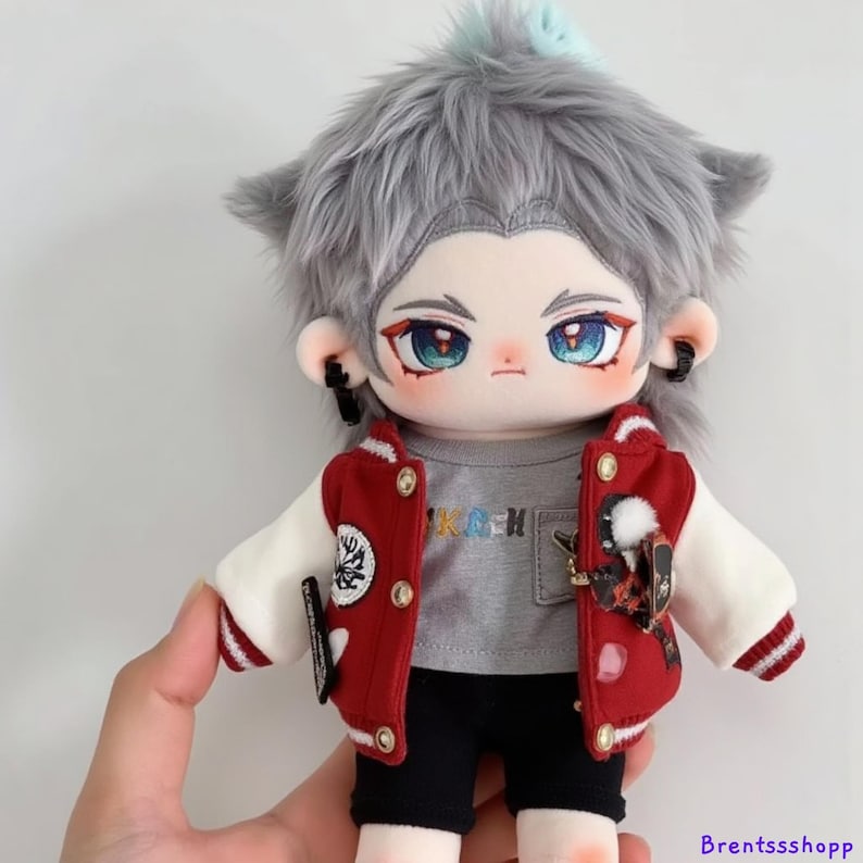 May include: A gray plush doll with a white and red varsity jacket, gray t-shirt with colorful text, black shorts, and gray hair. The doll has blue eyes and is wearing earrings.